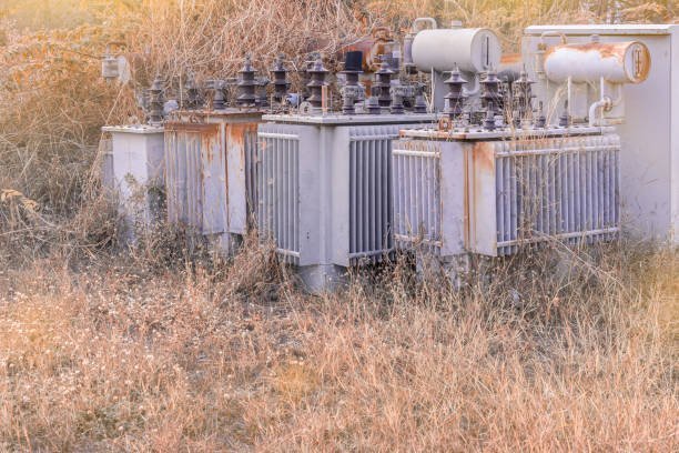 High-voltage power transformer outdoors with rust and weathering, situated in a grassy field, part of electrical infrastructure for power distribution.