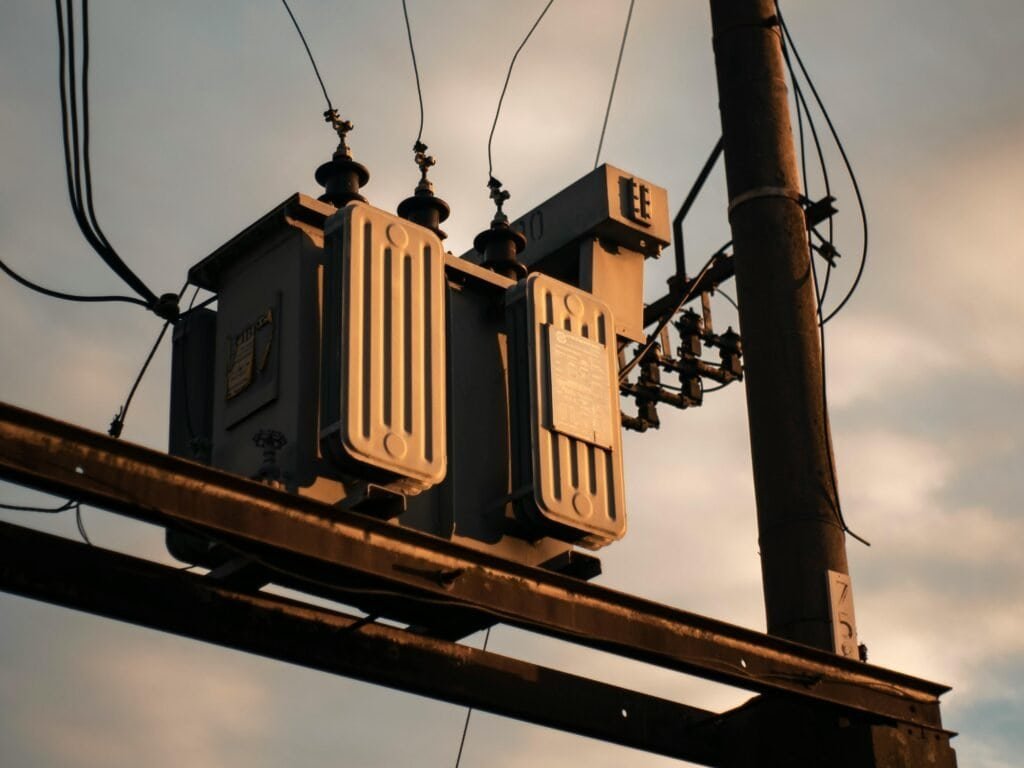 High-voltage electrical transformer mounted on utility pole, part of power distribution infrastructure, with overhead power lines against a sunset sky.