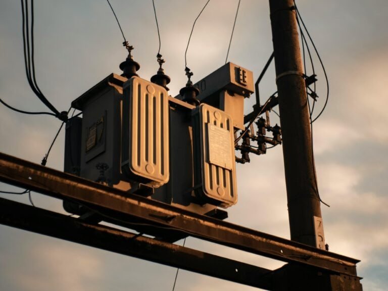 High-voltage electrical transformer mounted on utility pole, part of power distribution infrastructure, with overhead power lines against a sunset sky.