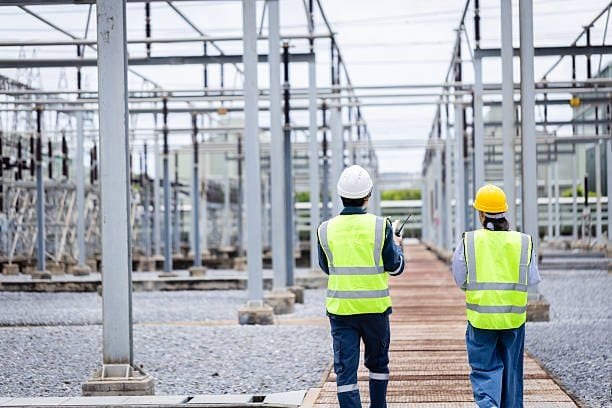 Two engineers in safety vests and helmets inspecting a large electrical substation or transformer facility.