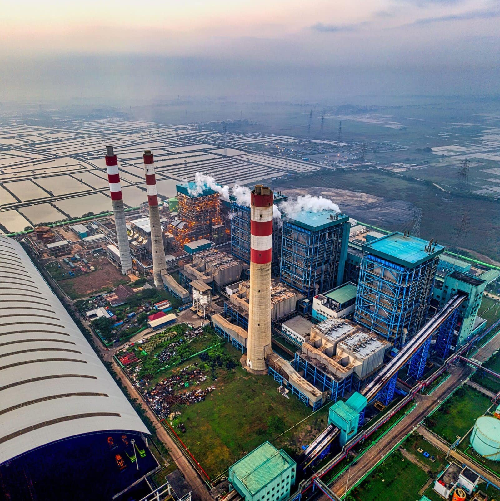 Aerial view of a large industrial power plant with multiple smokestacks emitting steam, surrounded by fields and infrastructure.