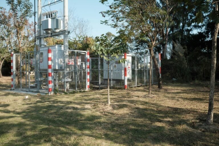 Electrical transformer station surrounded by trees and fencing, located outdoors in a grassy area.